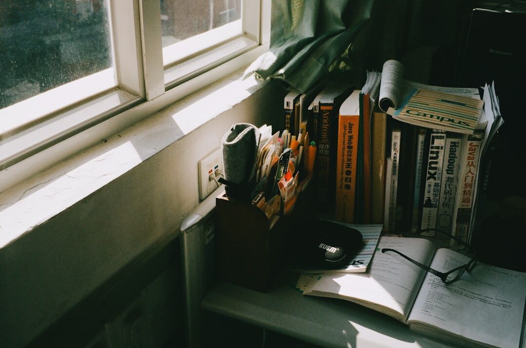 Small quiet home office with a wooden desk, lamp and stacked books near a window
